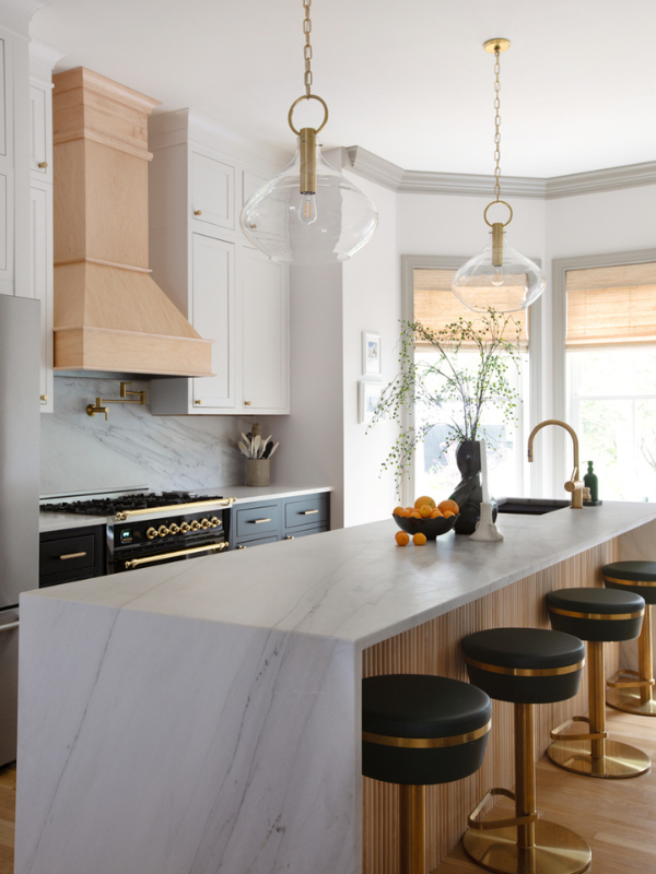 Stacked white inset shaker cabinets with black island cabinets, creamy waterfall countertops, funky glass and gold light fixtures, a natural wood kitchen hood