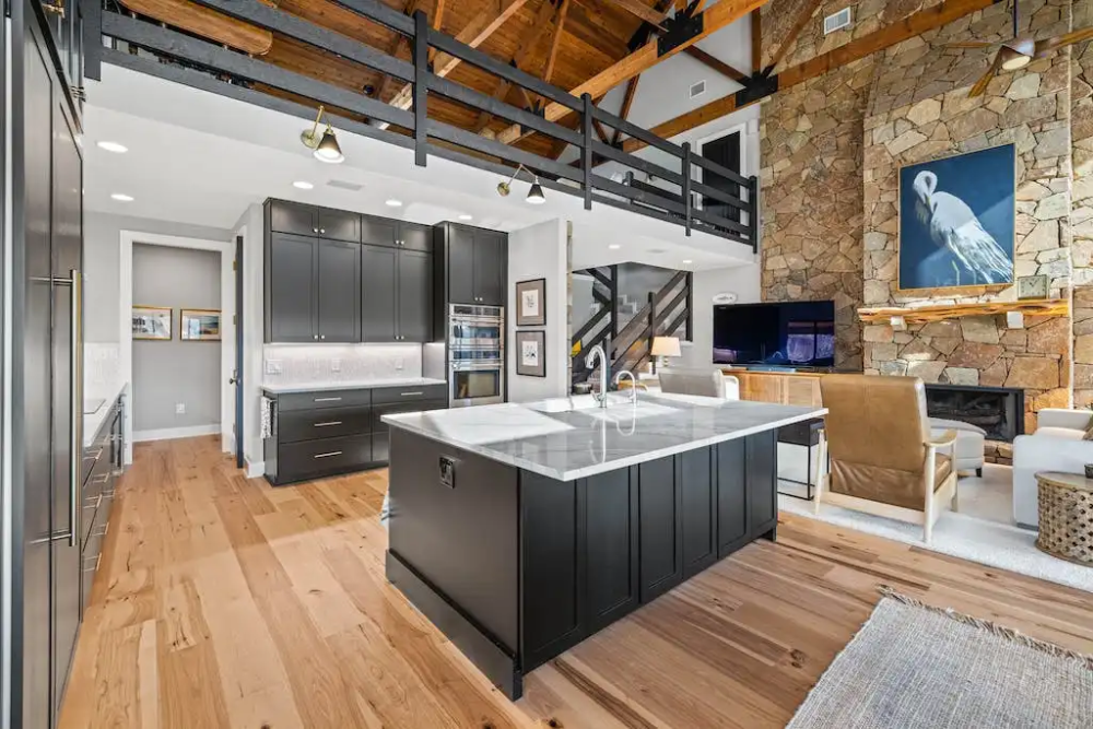 Stacked black shaker kitchen cabinets and island with white countertops and rustic wood floors by a rugged stone fireplace