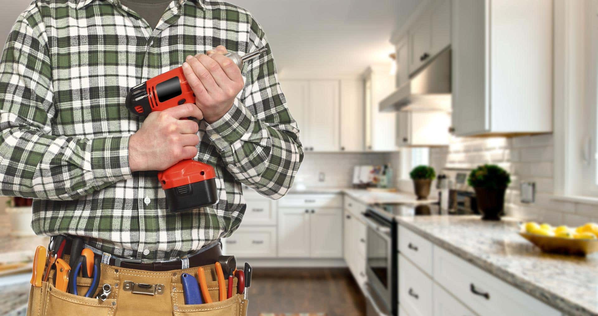 Professional contractor holding a power drill in a white shaker kitchen while wearing a tool belt.