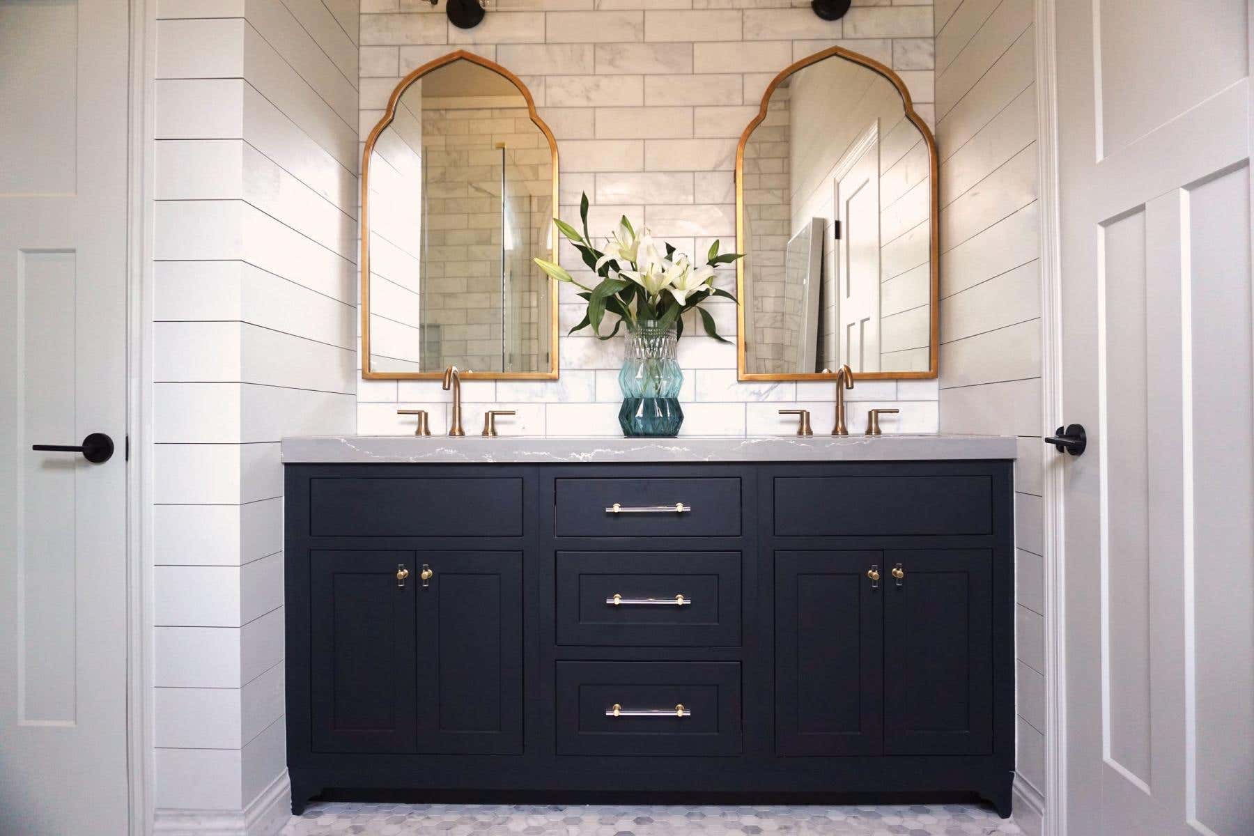 Black inset vanity cabinets with white countertops and marble subway tile surround and arched gold mirrors above both sinks