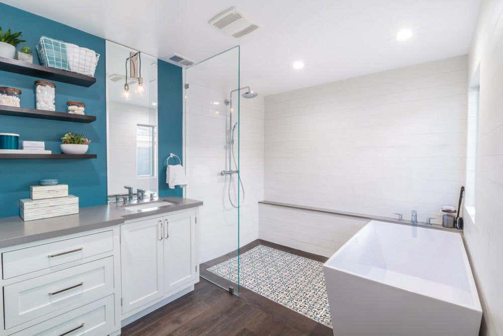 Modern white shaker bathroom cabinets with wood floating shelves next to glass shower and white free-standing tub