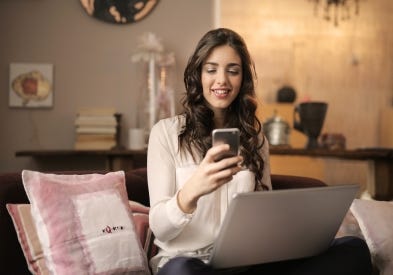 Woman sitting on her couch using her cell phone and computer