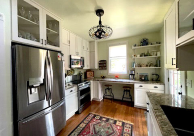 Stacked white kitchen cabinets with some glass inserts, floating wood shelves, a built-in desk, and farmhouse accents and lighting