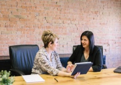 two women sitting at a table in front of a red brick wall going over a client presentation