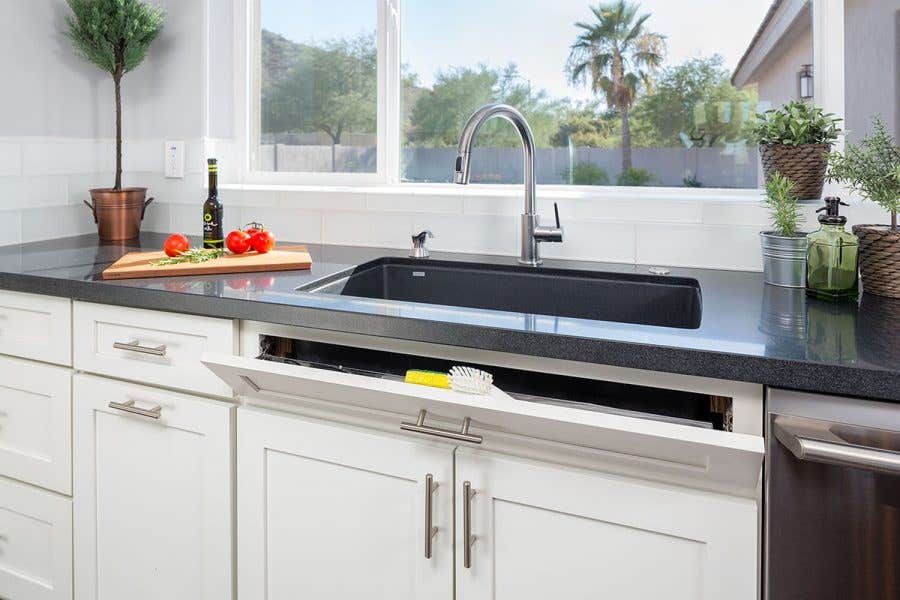 modern kitchen sink wall has large window, white shaker cabinets and sink tip out tray for small item storage and pull-out trash can.