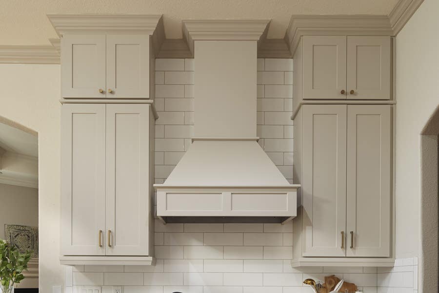 CliqStudios wood range hood in light gray paint called Urban Stone installed with tall wall cabinets on either side.