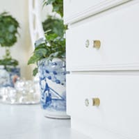 CliqStudios Bayport cabinets in White with glass knobs. Next to the cabinets is a plant in a blue and white china pot.