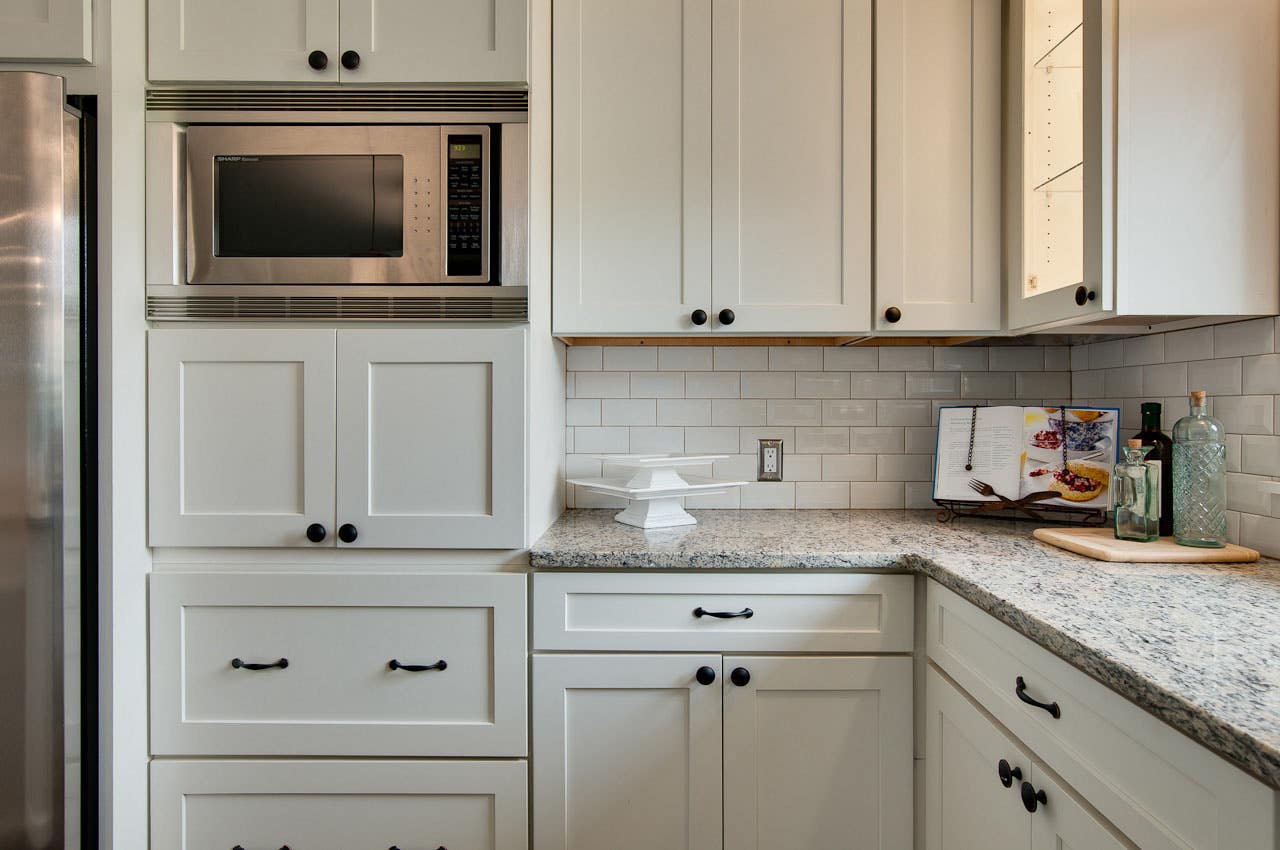 inside corner of kitchen has painted white cabinets in a shaker style with oiled brass pulls and knobs, granite countertop and built in microwave