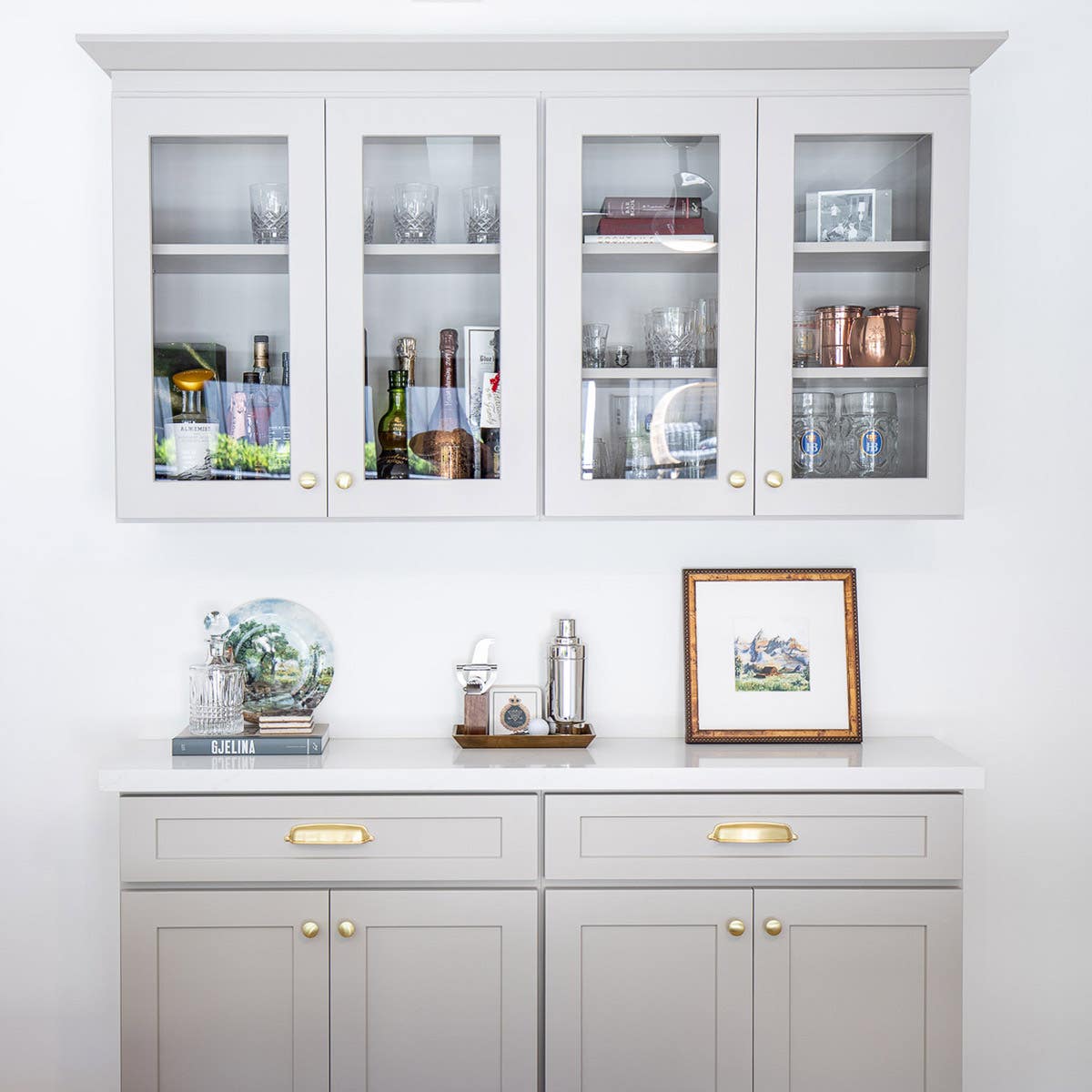 Taupe colored bar cabinetry with gold hardware and glass wall cabinet with crown molding above Taupe colored bar cabinetry with gold hardware and glass wall cabinet with crown molding above