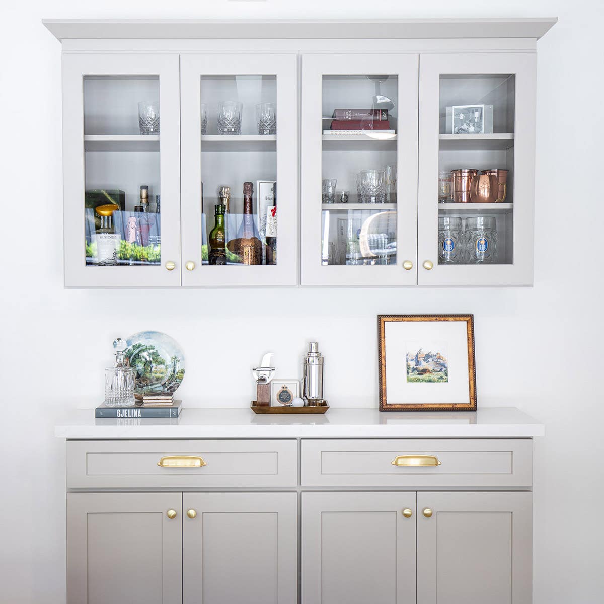 Taupe colored bar cabinetry with gold hardware and glass wall cabinet with crown molding above