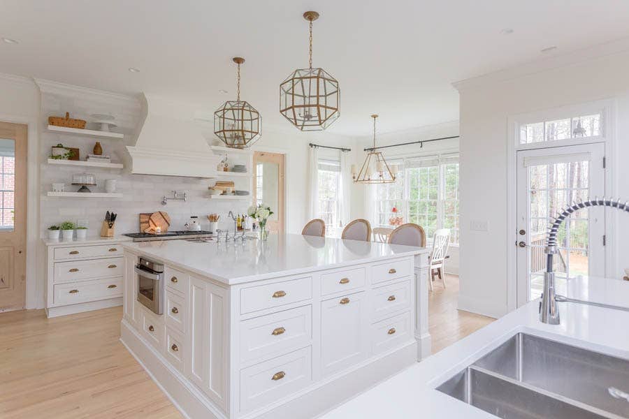 White inset shaker kitchen cabinets with large island, wood range hood, brass hardware, and floating shelves