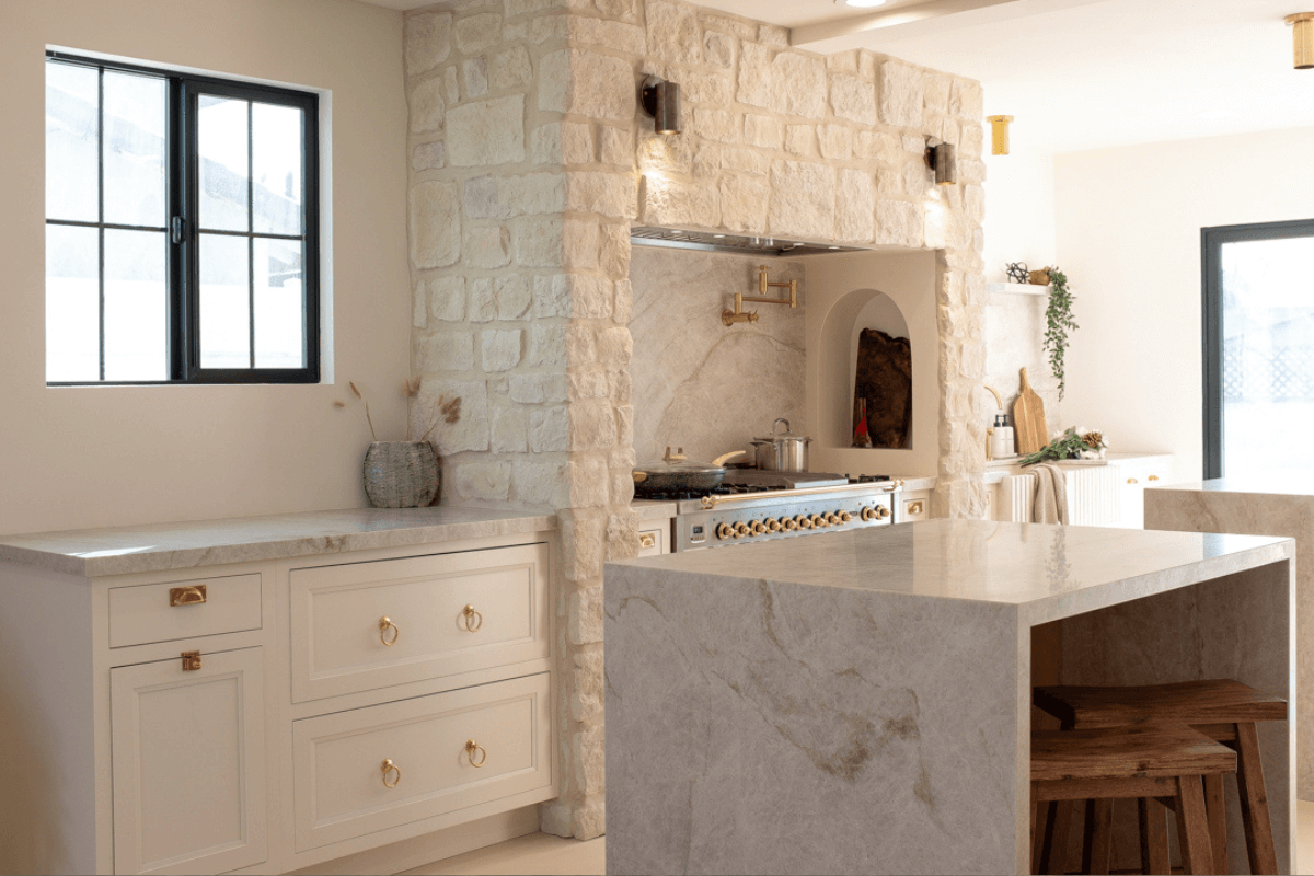 Mediterranean-style kitchen design with white inset shaker cabinets, limestone surround above the gas range and medium-tone inset kitchen island cabinets with waterfall countertops.