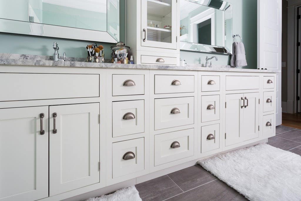 Deluxe new home features double vanity built from classic inset cabinets. Bathroom has dual custom mirrors, glass-front cabinet and drawer stacked on counter, and porcelain tile floor