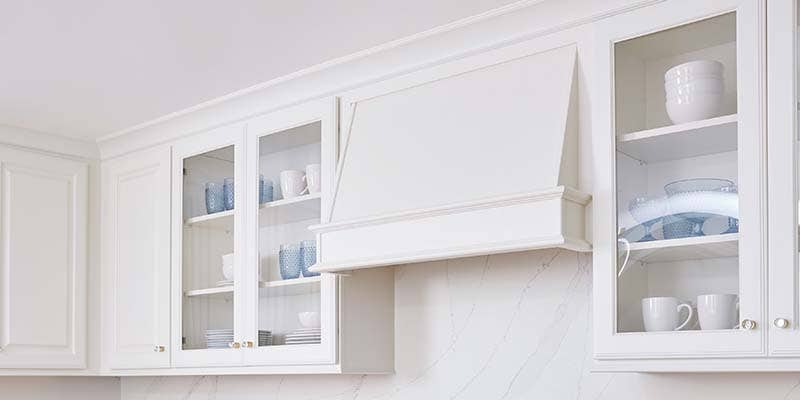 CliqStudios Decorative cabinet in Painted White with glass doors displaying glassware and dishware and a wood range hood in the Classic style CliqStudios Decorative cabinet in Painted White with glass doors displaying glassware and dishware and a wood range hood in the Classic style