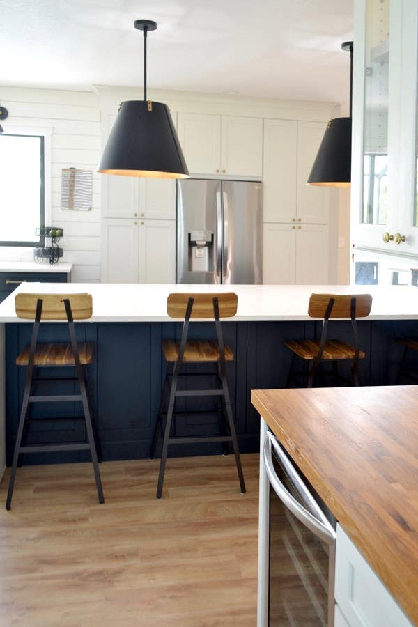 CliqStudios Shaker cabinets in black at the island and white for the wall cabinets. The island has a white waterfall countertop and tall bar chairs pulled up, with pendant lighting with black shades hanging above. Behind that is a wall of white cabinets,