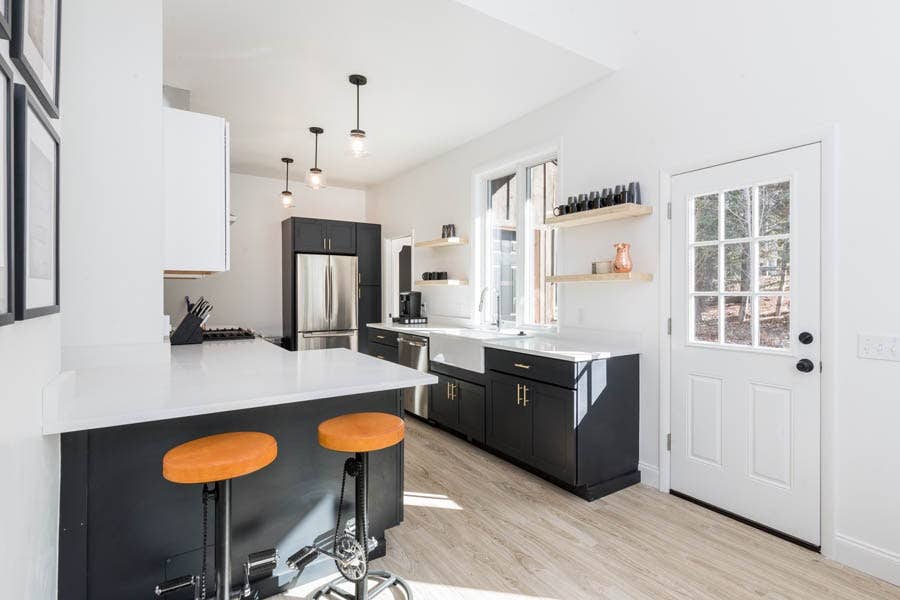Brightly lit white galley kitchen with black base cabinets, white apron sink under a window with floating wood shelves on both sides Brightly lit white galley kitchen with black base cabinets, white apron sink under a window with floating wood shelves on both sides