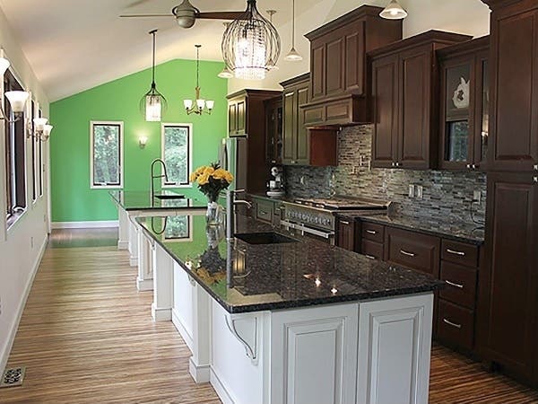 A wall of dark cherry cabinets with raised-panel doors faces two white kitchen islands and a bank of windows