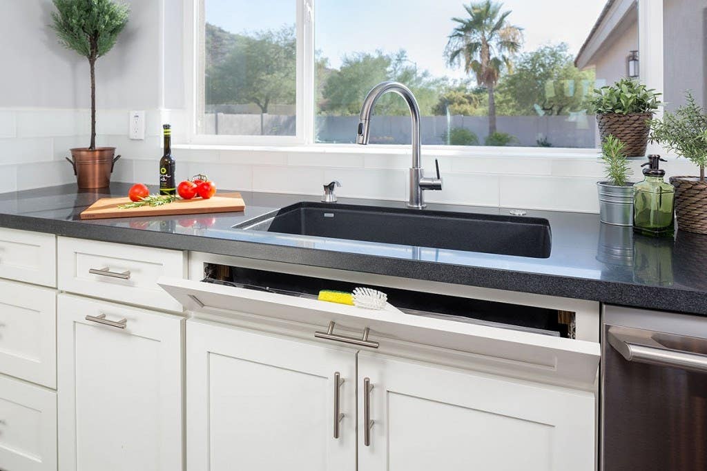 Modern kitchen sink wall has large window, white shaker cabinets and sink tip out tray for small item storage and pull-out trash can