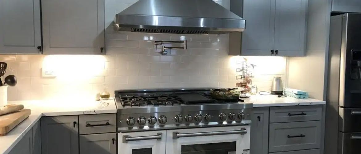 L-shaped kitchen layout with medium gray kitchen cabinets, white countertops and backsplash, black cabinet hardware, and a kitchen island featuring a large sink.