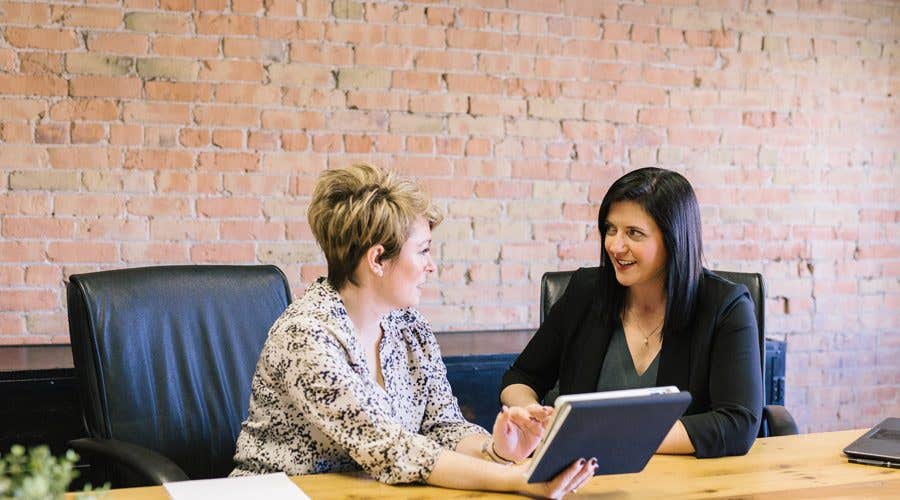 Two women sitting at a table in front of a red brick wall going over a client presentation