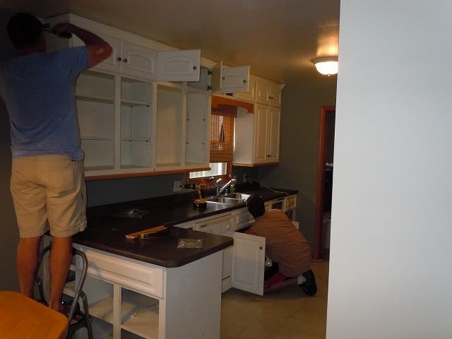 two men removing cabinet doors and disconnecting sink plumbing in kitchen with white cabinets and black countertops two men removing cabinet doors and disconnecting sink plumbing in kitchen with white cabinets and black countertops